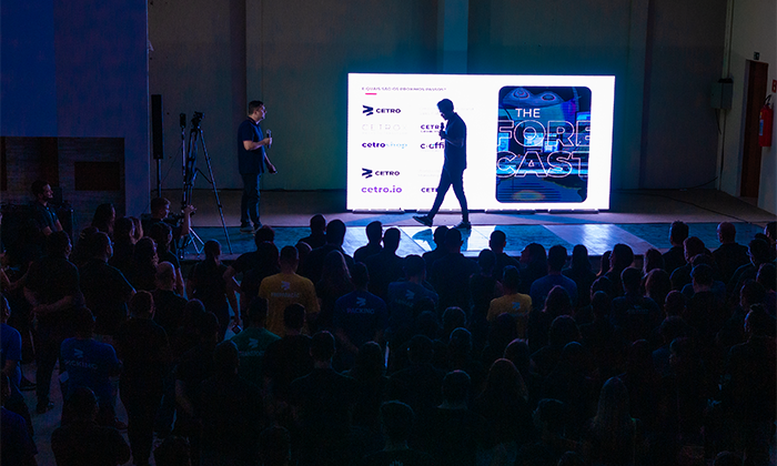 Foto em escala escura do CEO e fundador da empresa, Léo Mello, caminhando sobre um palco, com um painel digital exibindo uma luz branca em destaque ao fundo e colaborados da Cetro na plateia. usada para fala leo
