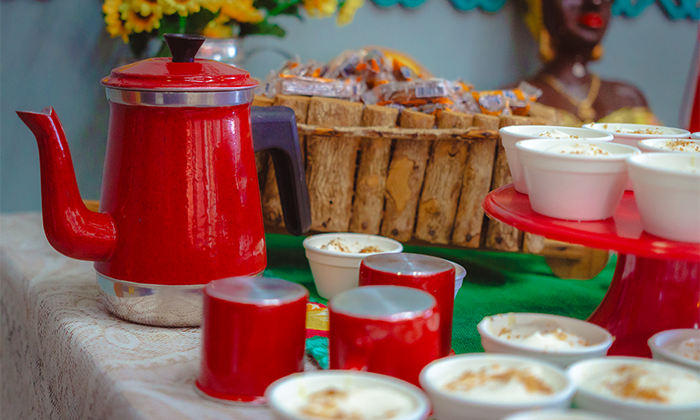 Fotografia de uma mesa posta do almoço mineiro na Cetro, com uma jarra de café e canecas de alumínio vermelhas, doces a base de amendoim e flores ao fundo.