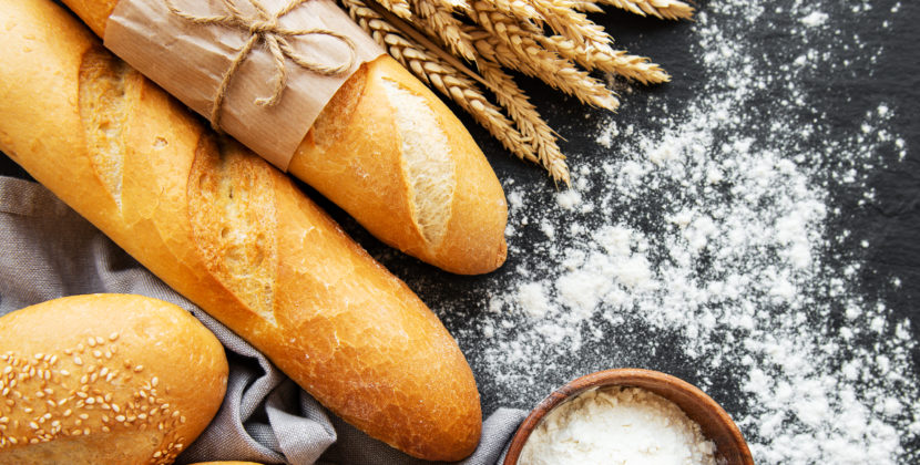Fotografia vista de cima de dois pães franceses, duas baguetes, ramos de trigo e uma cumbuca de farinha – ilustrando a diversidade do mercado de pães congelados.