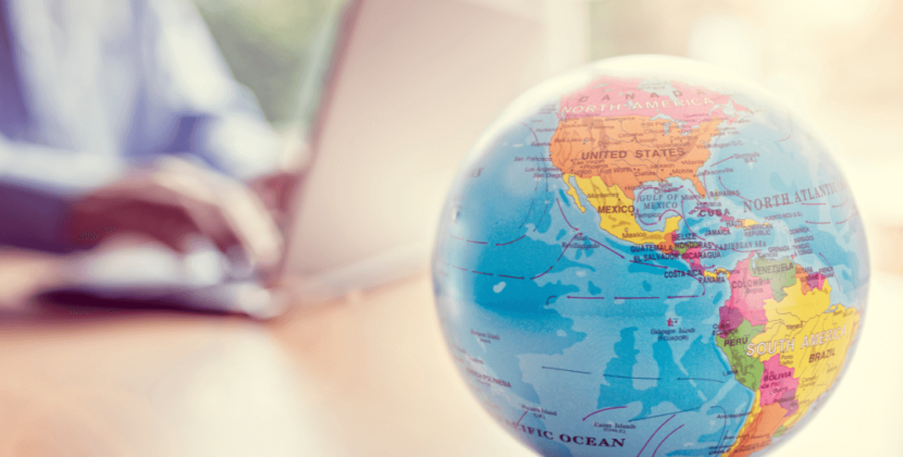 Foto de um globo em destaque, representando o processo de comércio no mercado internacional, com uma pessoa mexendo em um computador ao fundo.