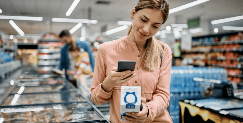 Imagem de uma mulher loira de pele branca escaneando uma embalagem sem rótulo com o celular na seção de frios de um supermercado.