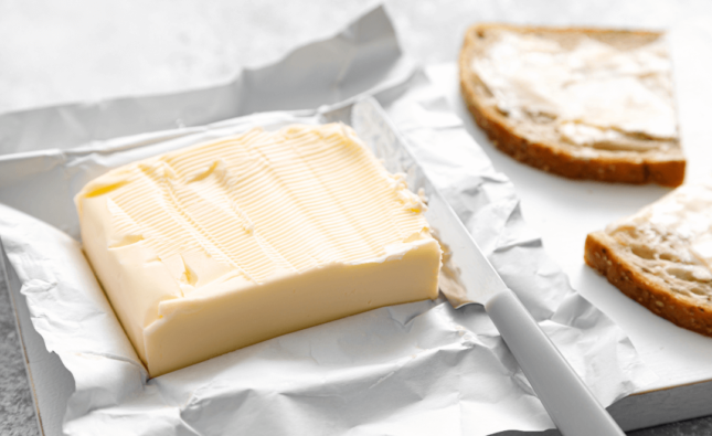 Foto de uma barra de manteiga sobre uma embalagem ao lado de uma faca e duas torradas com manteiga.