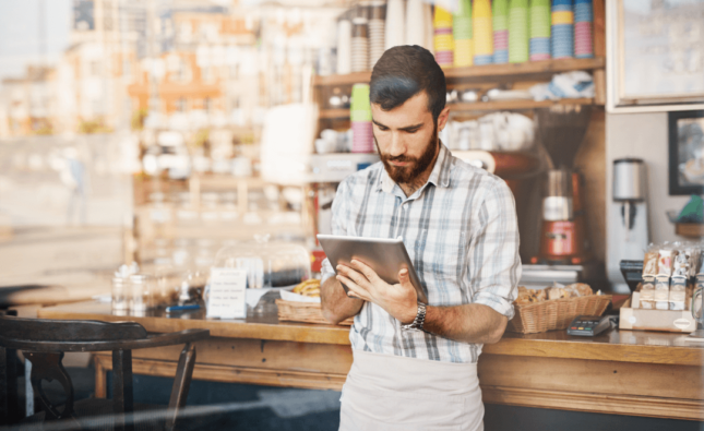 Imagem de um homem de avental segurando um tablet em uma cafeteria, representando o uso de ferramentas digitais para a comunicação efetiva com o cliente.