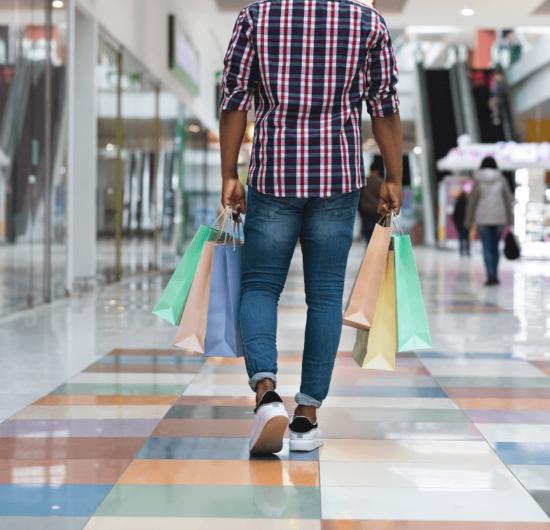 Imagem de um homem com sacolas de compra em um corretor de shopping, representando a alta da confiança do consumidor no mercado.
