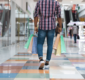 Imagem de um homem com sacolas de compra em um corretor de shopping, representando a alta da confiança do consumidor no mercado.