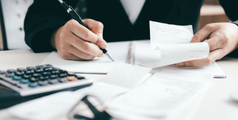 Imagem de um homem com um casaco social escrevendo sobre uma mesa cheia de papéis e uma calculadora, ilustrando o processo de declaração de rendimentos do MEI.