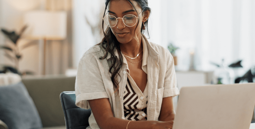 Imagem de uma mulher de óculos e roupas bege usando um notebook e olhando seu caderno de anotações, representando uma empreendedora fazendo o planejamento estratégico de sua empresa.