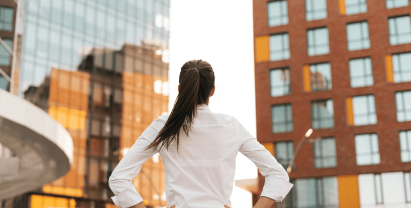 Imagem de uma mulher com o cabelo preso e camisa branca olhando para prédios comerciais com as mãos na cintura, representando o crescimento empresarial.