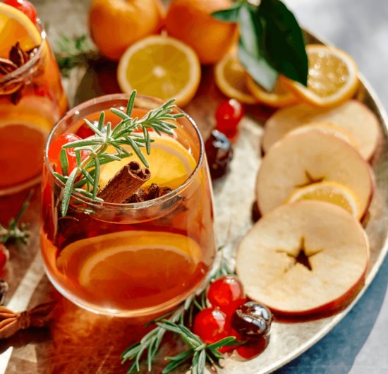 Fotografia de um drink laranja com infusão de frutas e especiarias, como rodelas de laranja, ramas de canela, cerejas e tomilho.