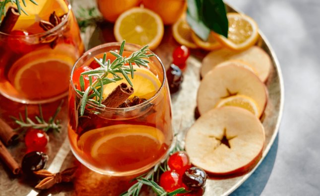 Fotografia de um drink laranja com infusão de frutas e especiarias, como rodelas de laranja, ramas de canela, cerejas e tomilho.
