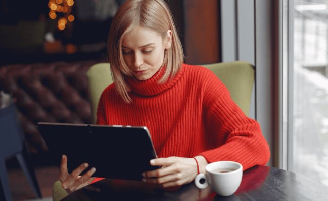 Imagem de uma mulher loira com suéter vermelho segurando um tablet sentada em uma mesa com uma xícara de café.