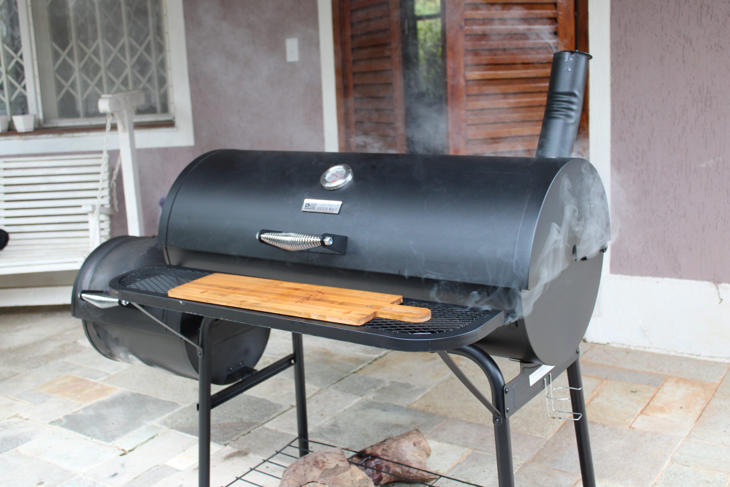 Fotografia do Defumador e Churrasqueira Charbroil Offser Smoker fechada, saindo fumaça de sua chaminé.