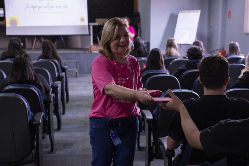 Fotografia dos colaboradores da Cetro assistindo à palestra sobre Outubro Rosa e das representantes do Grupo Amigas do Peito entregando folhetos aos participantes.