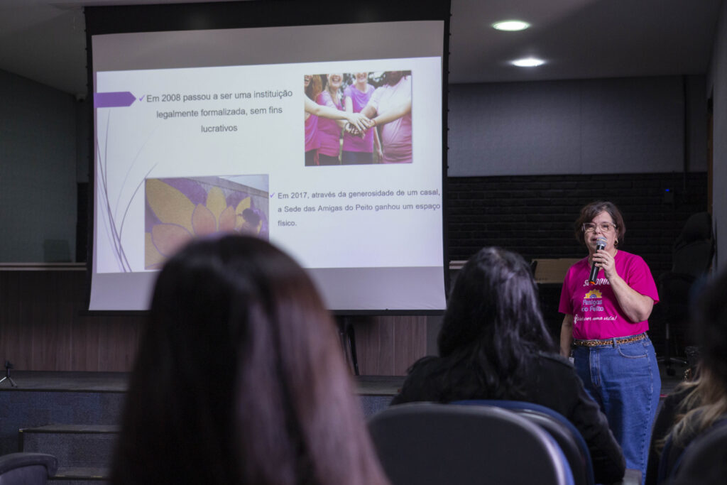Fotografia dos colaboradores da Cetro assistindo à palestra sobre Outubro Rosa e das representantes do Grupo Amigas do Peito entregando folhetos aos participantes.