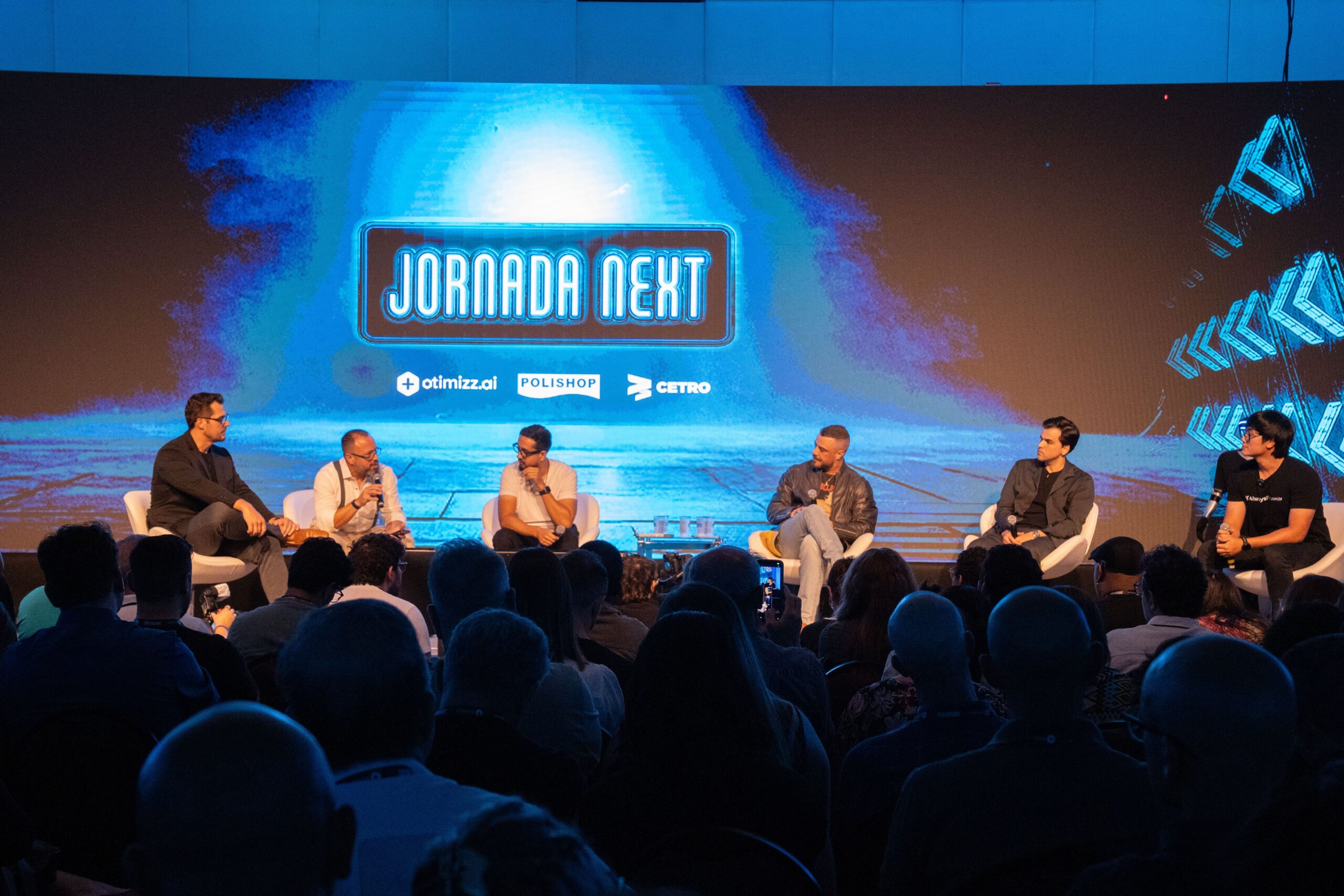 Fotografias dos palestrantes no palco e de um telão com a logo da Cetro e a palavra “patrocinador” no evento Jornada Next.