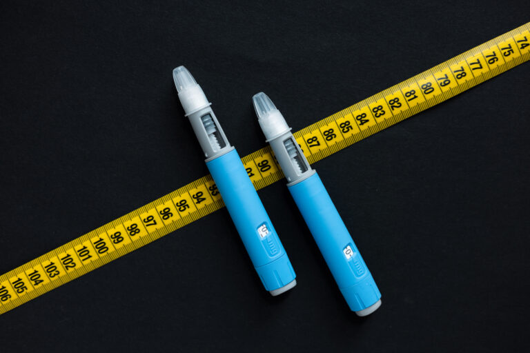 Imagem de duas canetas emagrecedoras azuis para perda de peso posicionadas sobre uma fita métrica amarela estendida sobre fundo preto.