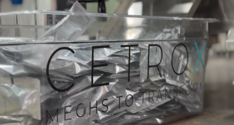 Fotografia de uma caixa transparente com a logo da CetroX contendo diversos sachês de cor prateada embalados por uma Empacotadora Automática de Sachês da marca.