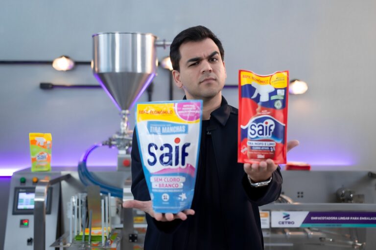 Léo Mello, CEO da Cetro, segurando duas embalagens stand-up pouch de produtos de limpeza em frente a uma Empacotadora Linear da Cetro.