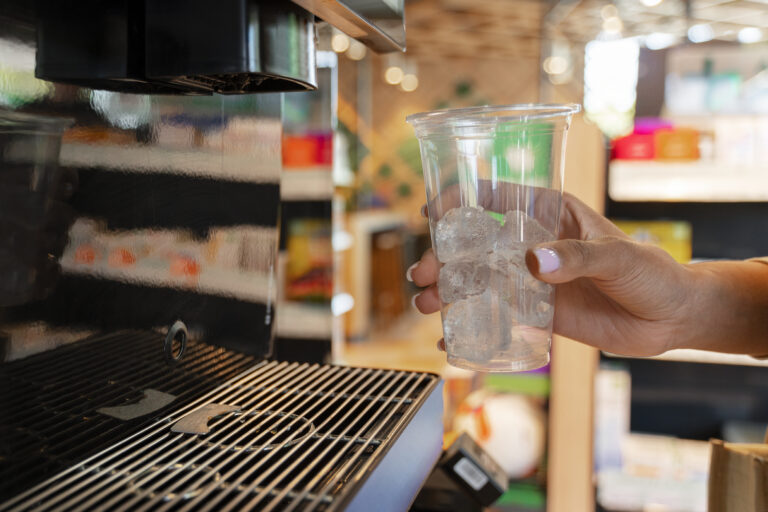Uma mão segurando um copo de plástico transparente com gelo ao lado de uma máquina de refrigerante.