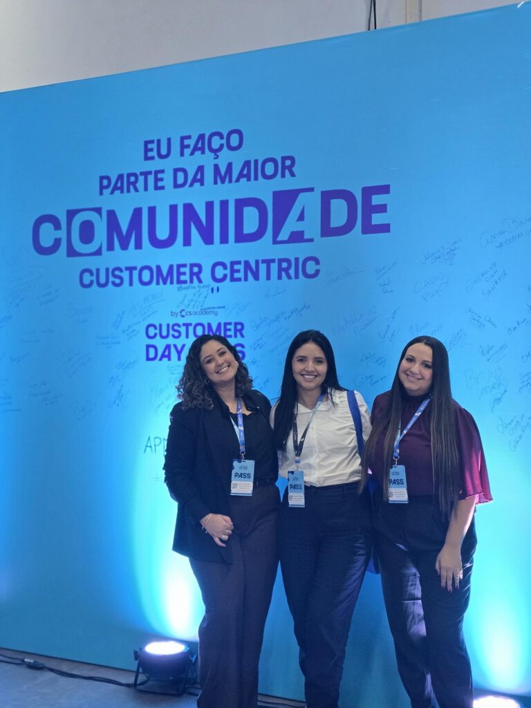 Fotografia do espaço do evento corporativo e das colaboradoras em frente a um painel personalizado com a logo do “Customer Day 2026”.