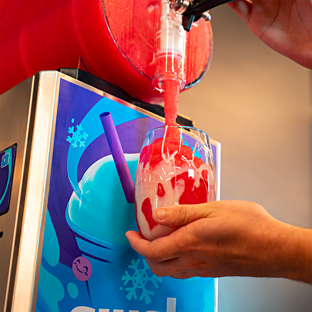 Máquina de Raspadinha e Bebidas Congeladas Slush e um copo de vidro com raspadinha vermelha decorada com calda, um morango e uma fatia de limão.