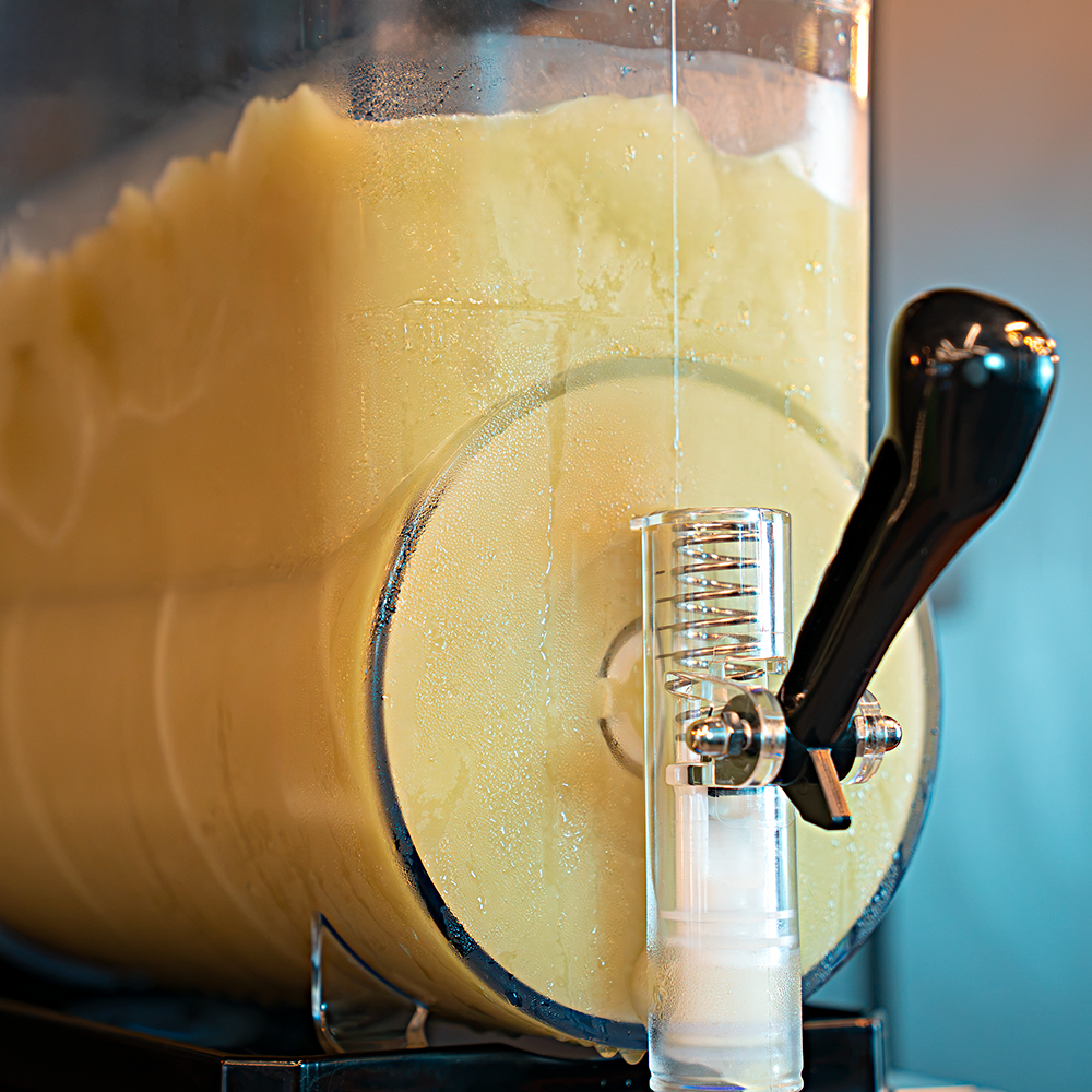 Uma Máquina de Raspadinha e Bebidas Congeladas Slush da Cetro Duty sendo abastecida com produto e uma raspadinha verde decorada com uma fatia de limão em um copo de plástico.