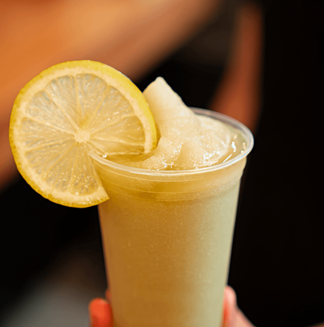 Uma Máquina de Raspadinha e Bebidas Congeladas Slush da Cetro Duty sendo abastecida com produto e uma raspadinha verde decorada com uma fatia de limão em um copo de plástico.