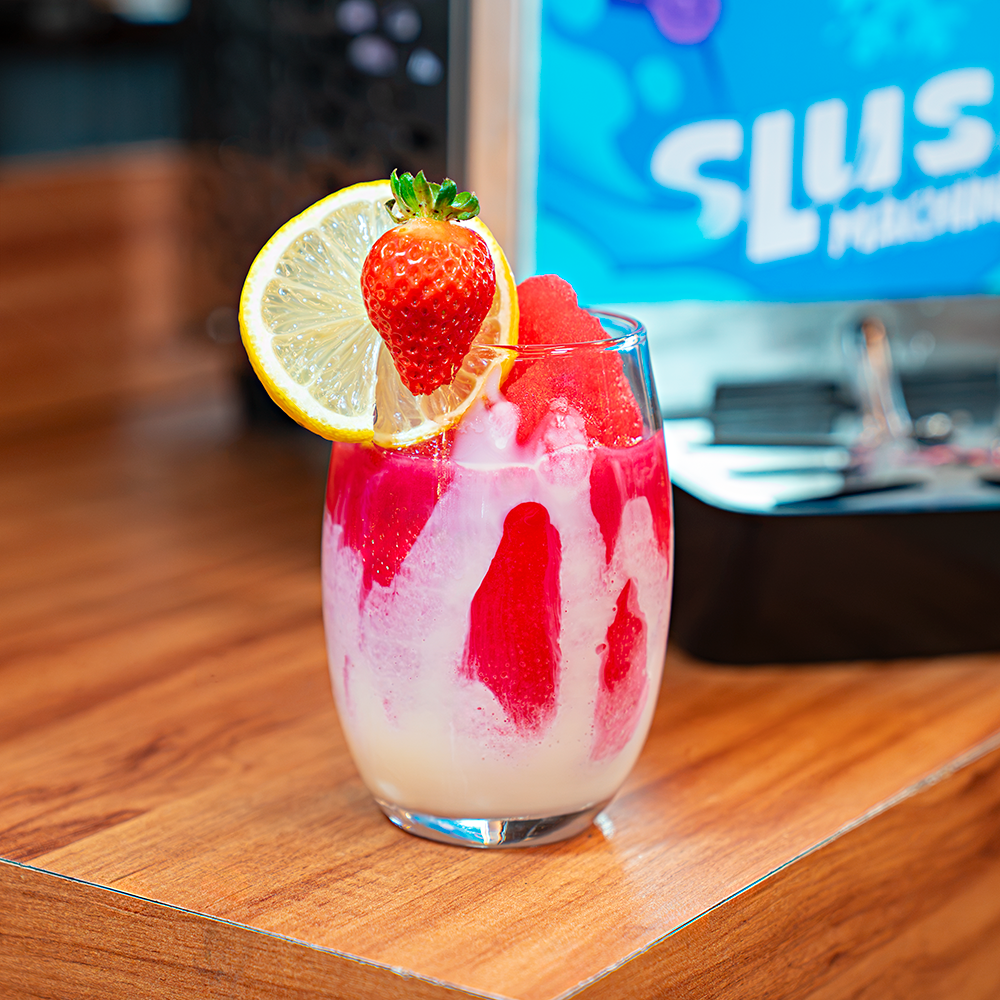 Máquina de Raspadinha e Bebidas Congeladas Slush e um copo de vidro com raspadinha vermelha decorada com calda, um morango e uma fatia de limão.